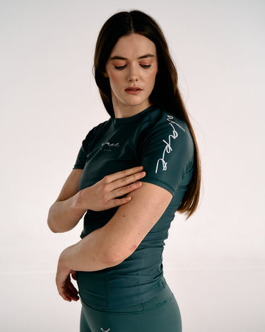 A woman modelling a dark teal short-sleeve BJJ rashguard with white script lettering on the sleeve, paired with matching dark teal athletic bottoms, against a plain light gray studio background.