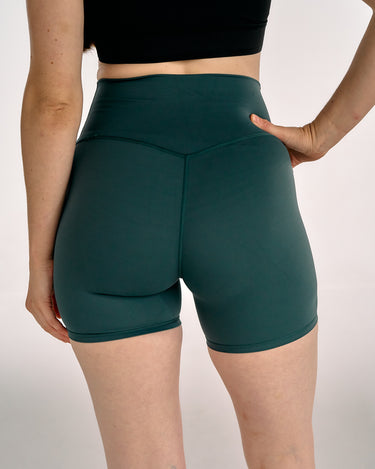 A close-up back view of a model wearing dark teal green high-waisted athletic shorts, paired with a black crop top, against a light gray studio background.