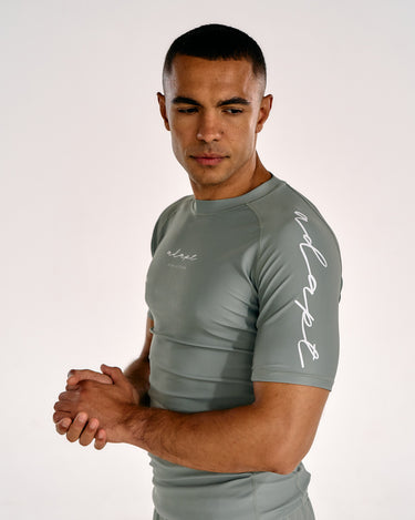 A man wearing a tight-fitting olive green athletic rash guard with white script logos on the chest and sleeve, posed against a plain light background.