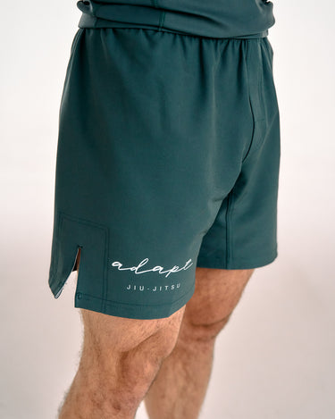 Dark teal green BJJ shorts with a side slit and white 'adapt JIU-JITSU' logo on the lower left leg, shown on a model against a light background.