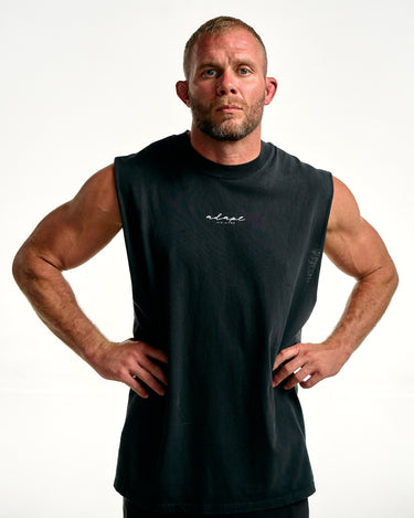 A muscular man with short blond hair and a beard poses with hands on hips wearing a black sleeveless athletic tank top with a white script logo on the chest, set against a plain white studio background.