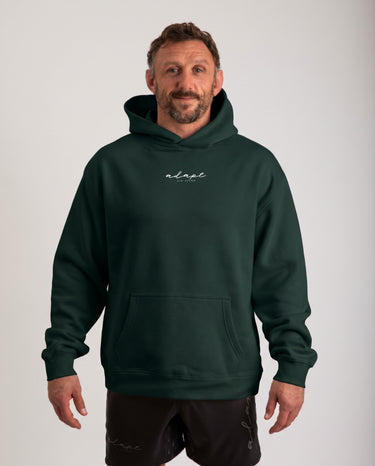 A man wearing a dark forest green pullover hoodie with a small white script logo on the chest, paired with black shorts, standing against a light gray studio background.