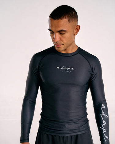 A man wearing a black long-sleeve BJJ rashguard with white script branding on the chest and sleeve, photographed against a plain light background.