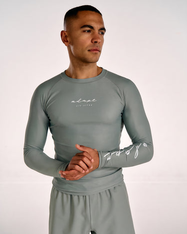 A man wearing a tight-fitting long-sleeve athletic shirt in a muted olive green color, featuring white script logos on the chest and left sleeve, paired with matching shorts against a plain light background.