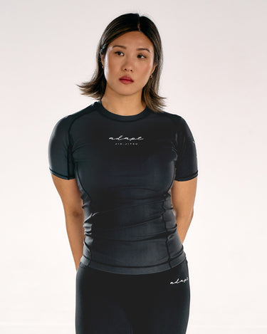 A woman modelling a black short-sleeve BJJ rashguard and matching black leggings with the 'adapt' brand logo, shot against a plain light background.