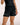A model wearing black BJJ shorts with a white 'adapt JIU-JITSU' logo on the left leg, paired with a black top against a light beige background.