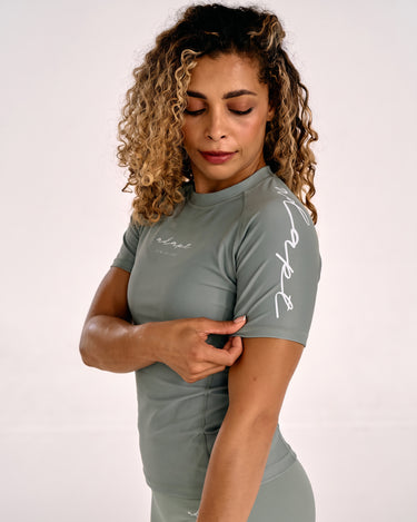 A woman with curly blonde hair wearing a olive green short-sleeve rashguard with white script lettering on the chest and sleeve, posing against a light gray studio background.