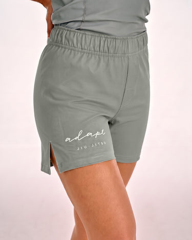 A close-up of a person wearing olive green BJJ training shorts with a side slit and a white 'adapt JIU-JITSU' logo printed on the left leg, against a light pink studio background.