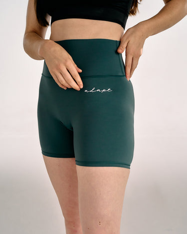 A model wearing teal high-waisted BJJ compression shorts with a side pocket and the brand name 'adapt' embroidered in white on the front, paired with a black sports bra against a light gray studio background.
