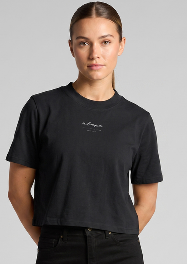 A woman wearing a black cropped t-shirt with a small white script logo on the chest, paired with black jeans, against a solid gray studio background.