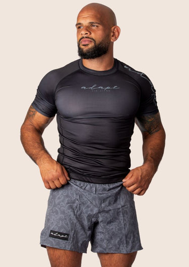 A man wearing a black Adapt Classic Rashguard with short sleeves and paired with gray paisley shorts.