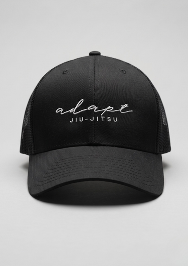 A black trucker-style baseball cap with white embroidered text reading 'adapt JIU-JITSU' on the front, shown against a light gray studio background.