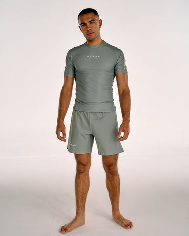 A man modeling a matching short-sleeve rash guard and swim shorts set in a muted olive green color, standing against a plain white studio background.
