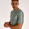 A man wearing a tight-fitting olive green athletic rash guard with white script logos on the chest and sleeve, posed against a plain light background.