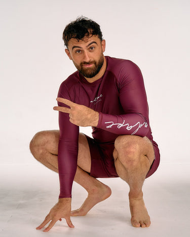A man wearing a tight-fitting maroon long-sleeve rash guard and matching shorts, crouching in a studio setting and pointing to the script logo on his sleeve.