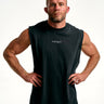 A muscular man with short blond hair and a beard poses with hands on hips wearing a black sleeveless athletic tank top with a white script logo on the chest, set against a plain white studio background.