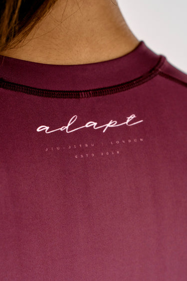 Close-up of a person wearing a maroon BJJ rashguard crewneck shirt with 'adapt' printed in white script on the chest, with 'Jiu-Jitsu London Est. 2018' printed below.