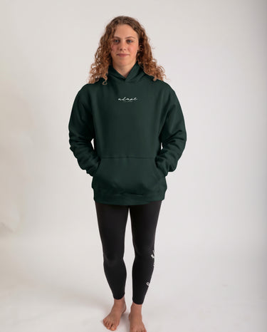 A woman with curly light brown hair stands barefoot against a plain white studio background, wearing a dark forest green pullover hoodie with a small white script logo on the left chest and black leggings.