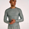 A man wearing a tight-fitting long-sleeve athletic shirt in a muted olive green color, featuring white script logos on the chest and left sleeve, paired with matching shorts against a plain light background.