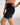 A close-up of a model wearing black high-waisted BJJ compression shorts with a small white script logo on the left hip, set against a plain light gray studio background.