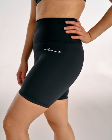 A close-up of a model wearing black high-waisted BJJ compression shorts with a small white script logo on the left hip, set against a plain light gray studio background.