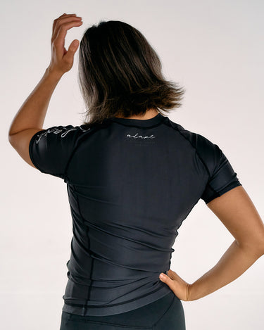 A woman shown from behind wearing a fitted black BJJ short-sleeve rashguard with white script branding on the back and shoulders, posed against a plain light background.