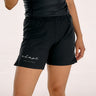 A model wearing black BJJ shorts with a white 'adapt JIU-JITSU' logo on the left leg, paired with a black top against a light beige background.