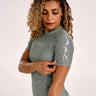 A woman with curly blonde hair wearing a olive green short-sleeve rashguard with white script lettering on the chest and sleeve, posing against a light gray studio background.