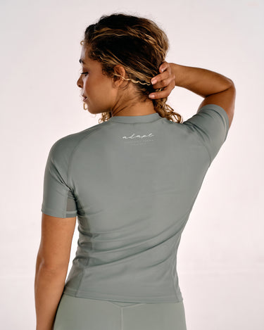 A woman shown from the back wearing a fitted short-sleeve BJJ rashguard top in a olive green color. The top has a small white script logo on the upper back.