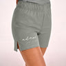A close-up of a person wearing olive green BJJ training shorts with a side slit and a white 'adapt JIU-JITSU' logo printed on the left leg, against a light pink studio background.