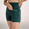 A model wearing teal high-waisted BJJ compression shorts with a side pocket and the brand name 'adapt' embroidered in white on the front, paired with a black sports bra against a light gray studio background.