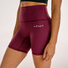 A close-up of a model wearing high-waisted BJJ compression shorts in maroon with a small white script logo on the front, paired with a black sports bra against a plain light background.
