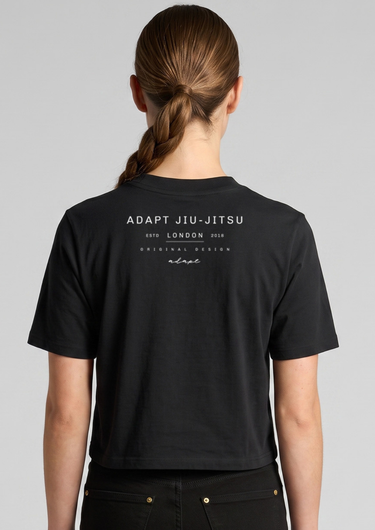 A woman viewed from the back wearing a black short-sleeve crop top T-shirt with white printed text reading 'ADAPT JIU-JITSU' and 'ESTD LONDON 2018' on the upper back.