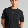 A woman wearing a black cropped t-shirt with a small white script logo on the chest, paired with black jeans, against a solid gray studio background.